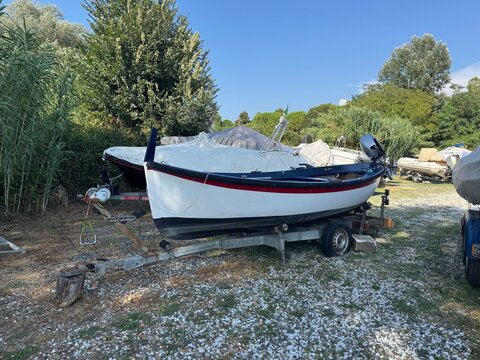 Classic small fishing boat on trailer parked in outdoor boatyard with trees under blue sky. Marine vessel storage concept highlights boat maintenance and nautical equipment.