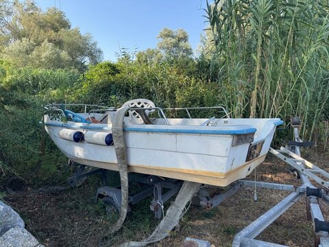 Small motorboat parked on trailer in outdoor overgrown storage area with trees and reeds under blue sky. Abandoned vessel concept represents boat maintenance and maritime storage.