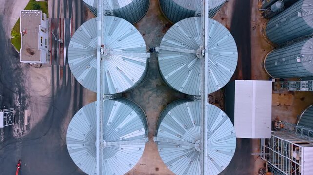 Top down aerial shot of circular metal grain silos. Four large silver storage tanks with geometric tops form a symmetrical pattern from above.