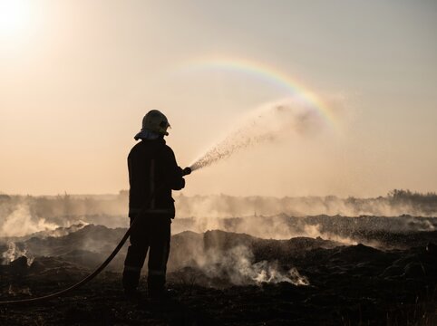 Brave firefighter in heavy protective gear sprays water from a long hose to extinguish burning land during a sunny afternoon while creating a bright vibrant rainbow in the misty air.