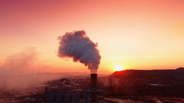 Industrial smokestack releasing smoke plume at sunset. The setting sun behind hills illuminates thick smoke rising from a factory chimney into an orange sky.
