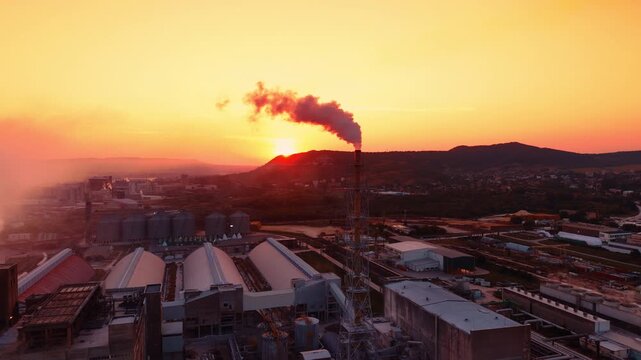 Industrial complex with smoke emitting chimney at sunset. Red smoke rises from a central factory chimney as the sun sets over hills behind large industrial buildings.