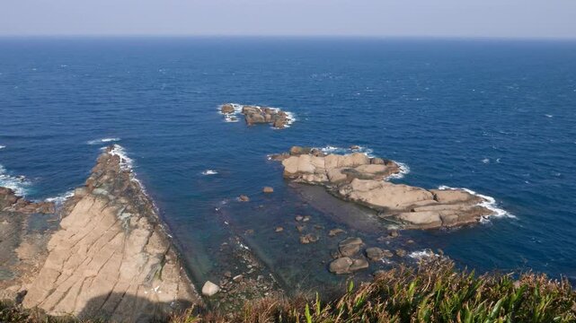 Vibrant blue sea waters from end of Yehliu Geopark cape, rock ledge sink against shore. Strong breeze from sea, green grass blades flutter on wind. Washed stone shelf break around, high angle shot.