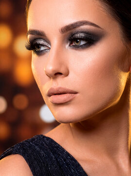 Glamorous beauty portrait of a woman with dark silver smokey eye makeup and nude lips against a warm golden bokeh background