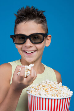Excited boy wearing 3D glasses holding bucket of popcorn isolated on blue background. Happy kid watching movie in cinema studio portrait