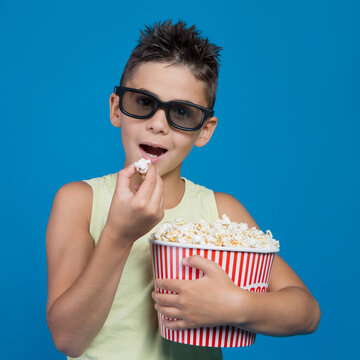 Boy wearing 3D glasses eating popcorn from bucket isolated on blue background. Happy child watching movie in cinema entertainment concept