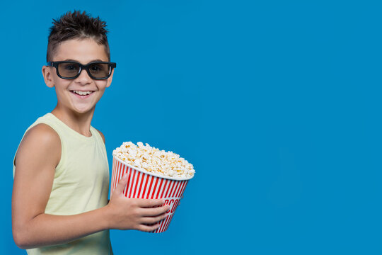 Happy boy wearing 3D glasses holding popcorn bucket with copy space isolated on blue background. Smiling kid watching movie in cinema banner concept