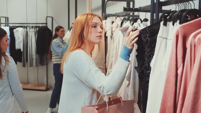 Young woman examines black dress on hanger in fashion store, shopping for stylish outfits and making wardrobe choice during leisure