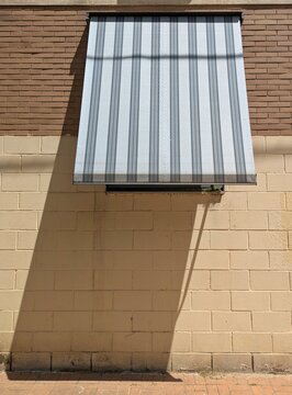 Toldo de tela rayada instalado para proteger una ventana de un edificio de los rayos de sol en verano, proyectando su sombra sobre la fachada y el suelo 