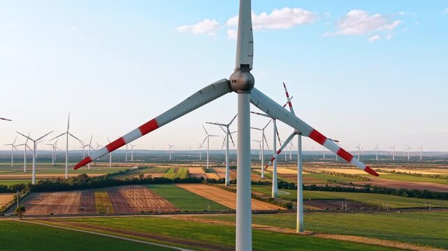 Close aerial view of wind turbine rotor and blades over farmland. Red tipped propeller blades frame a detailed panorama of rectangular cultivated fields below.