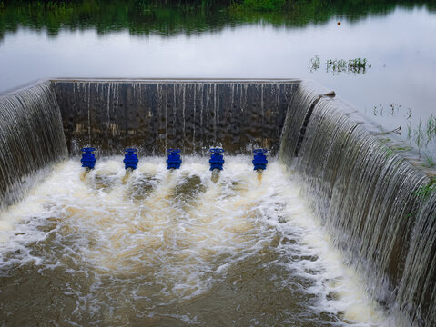 A concrete weir structure controls water flow in a rural river. The storage system features blue valves releasing stream current during irrigation supply management for agriculture.