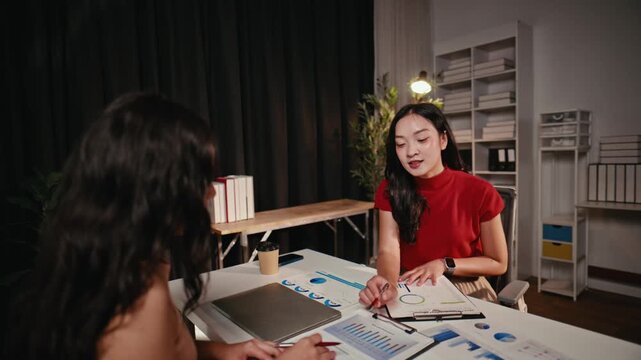 Two young professional Asian women discussing business strategy and analyzing financial charts during a meeting in the office at night