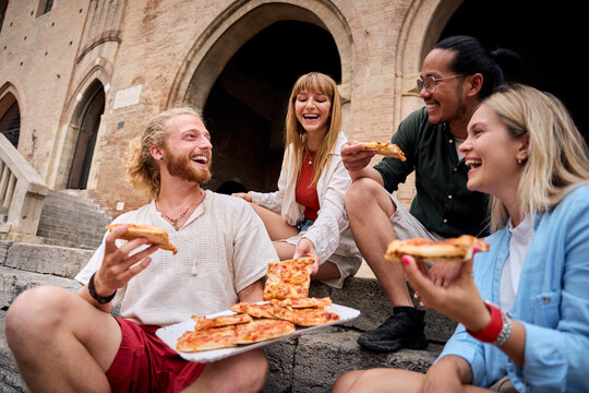 Group of cheerful young multi-ethnic friends enjoying pizza slices sitting on ancient stone steps during their summer vacation, having fun together