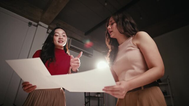 Two young asian businesswomen enthusiastically brainstorming and discussing project plans from papers while working late at the office