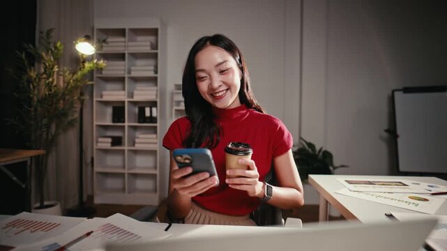 Smiling young Asian woman holding a coffee cup and texting on her mobile phone. She is working late in a modern office at her desk