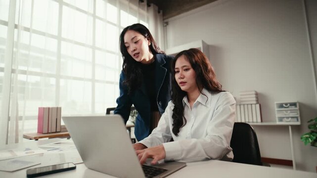 Two young asian businesswomen collaborating in a modern office. A manager is guiding her colleague while she works on a laptop computer