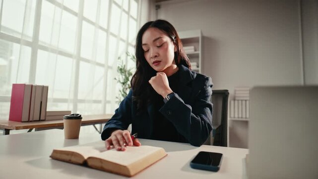 Serious young asian businesswoman sitting at her desk, thinking deeply while reading a book. Concentrated female student preparing for an exam