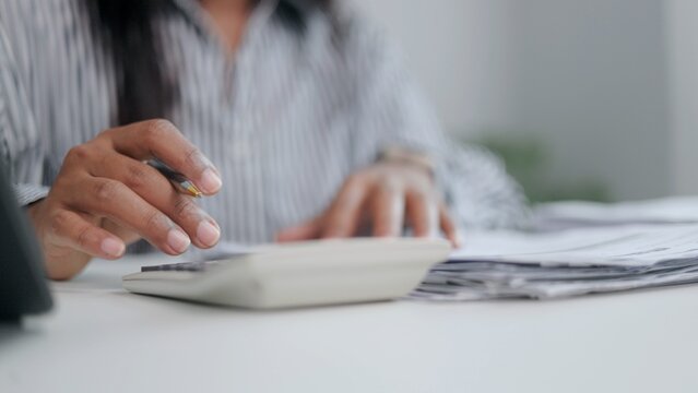 A close up view of an Asian female accountant typing on calculator keys while analyzing corporate expense sheets and financial statement paperwork at office desk workspace.