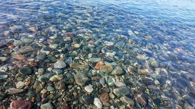 Slow motion clear sea water gently flows over colorful smooth pebbles on shallow shore.