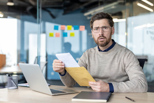 Businessman sitting at an office desk, looking surprised and worried while opening a brown envelope and reading a letter, feeling stress and anxiety about the unexpected message