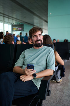 Smiling man sitting using phone while waiting for his flight at the airport departure area, looking the views. Vertical image with copy space.
