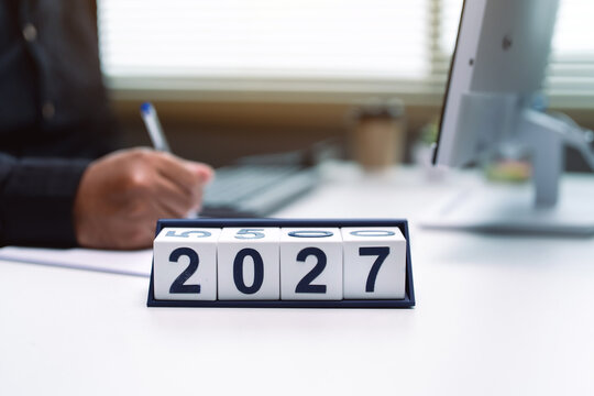 2027 year block calendar on desk with blurred businessman writing in modern office background, business planning and future strategy concept