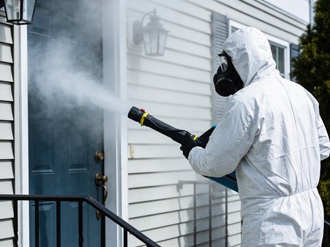 A person in a protective suit and gas mask uses a sprayer to disinfect a suburban home front door.