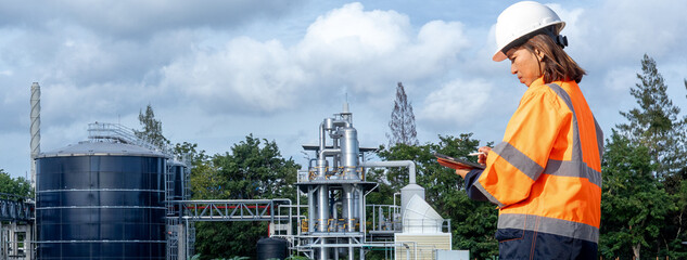 A woman wearing a helmet and bright safety gear stands at a factory site. She uses a tablet to check information while machinery and storage tanks are in view under a cloudy sky © Happy Photo