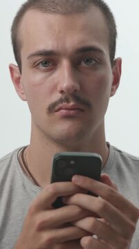 Young man holding a smartphone against a white background, typing, looking up directly at the camera with a calm expression, studio portrait.