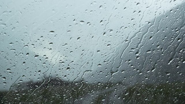Raindrops dripping down a window pane with a blurred mountain landscape in the background. Commercial stock video footage for bad weather, rainy season, melancholy, and cozy indoor concepts.