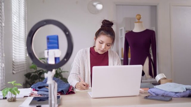 Young Asian fashion entrepreneur taking notes while filming online clothing business video with ring light in studio