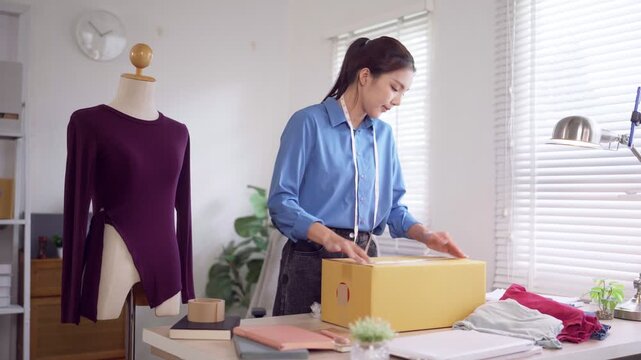 Young Asian fashion entrepreneur sealing yellow shipping box for online clothing order in bright home studio workspace