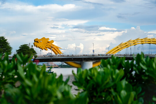 The striking head golden dragon head of Da Nang's Dragon Bridge stands over the Han River, flanked by green trees and decorative red lanterns under a cloudy sky