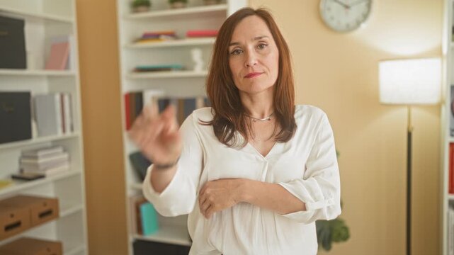 Woman pinches thumb and forefinger to show a small amount, arms folded, in a studio office with bookshelves and clock; precision contemplation.