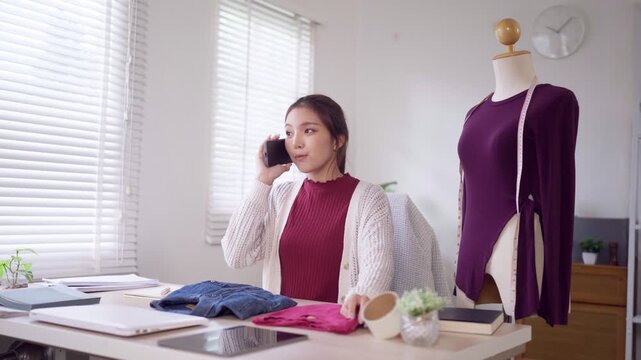 Young Asian fashion designer taking phone order while reviewing clothing inventory in bright home studio