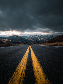 A long, empty asphalt road stretches into the distance toward dark mountains under a dramatic sky, featuring bright yellow center lines in a minimalist composition.