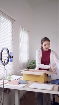 Young Asian online seller unpacking clothing inventory from cardboard box in bright home office workspace