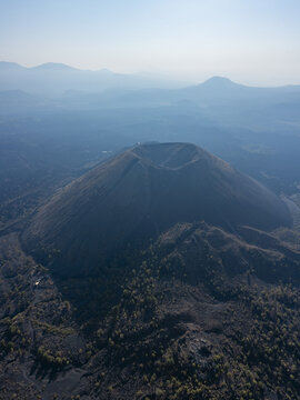 Aerial view of the Paricutin Volcano crater surrounded by vast volcanic lava fields and distant mountains under a hazy sky in Paricutin, Michoacan, Mexico.