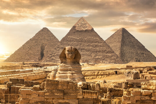 Famous Giza Pyramids stand behind the Great Sphinx under a dramatic sunset desert sky, Egypt.