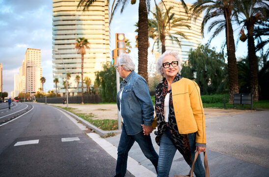 Senior couple walking hand in hand across a city street at dusk. Candid urban scene with modern buildings and palm trees, conveying companionship, active aging and independence.