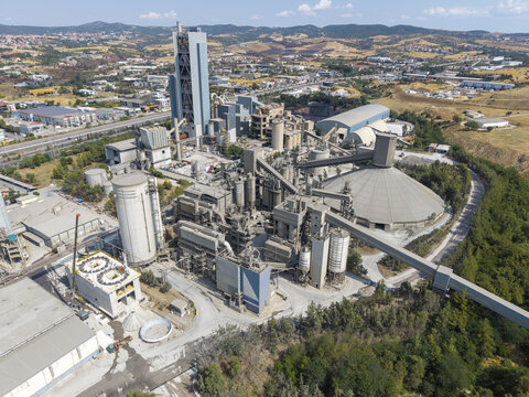 Aerial view of the TITAN Cement factory complex featuring industrial silos, conveyor belts, and processing structures surrounded by hills and roads in Thessaloniki, Central Macedonia, Greece.