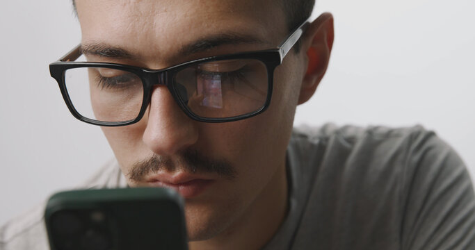 Focused young man with glasses looking down at his smartphone screen on a white studio background.