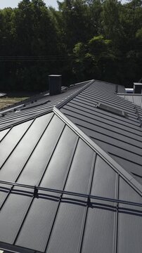 Vertical video. Aerial view moving along the black metal tile roof of a modern house under construction, detailing the material, gutter system, and snow guards on a sunny day