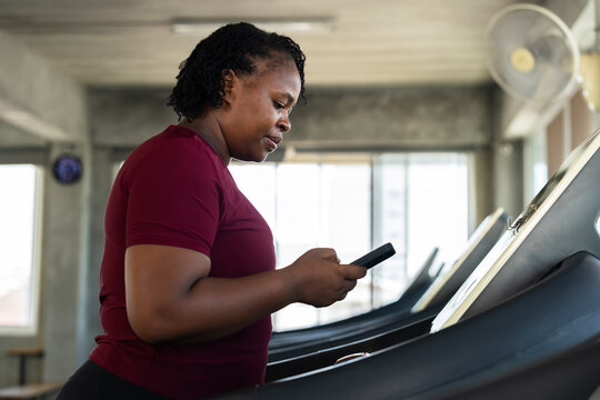 Plus size African American woman checking phone while standing beside treadmill in modern gym, active fitness lifestyle photo for cardio training, wellness routine, body confidence and energy.