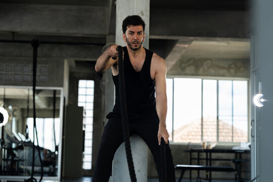 Adult Caucasian man training with battle rope in modern gym, determined male athlete doing functional strength endurance workout, high intensity fitness routine, personal training content for gym ads