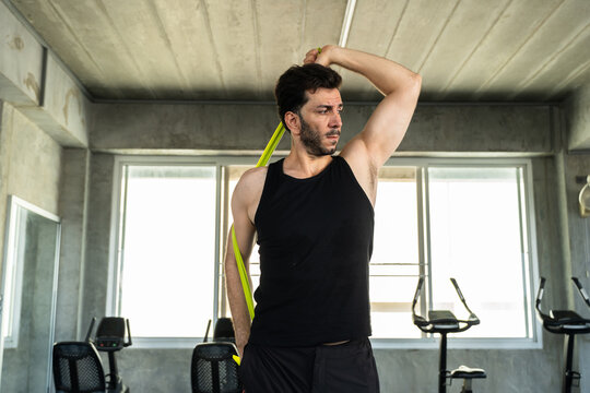 Adult Caucasian man stretching shoulder with green resistance band in modern gym, mobility warm up before strength workout, active male athlete fitness lifestyle for personal training and wellness ads