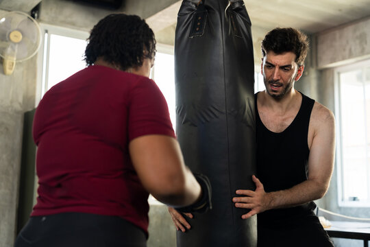 Plus size African American woman wearing boxing gloves trains with male instructor holding punching bag in modern gym, cardio boxing practice, personal coaching, partner workout motivation, teamwork.