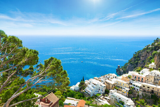 Positano hillside rises above blue Tyrrhenian Sea with white buildings, cliffs, pines, and bright sky. Positano coastline stretches toward calm horizon under clear Mediterranean daylight.