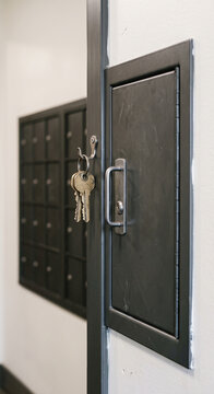 Vertical close-up of keys hanging from the lock of an open dark metal mailbox panel with compartment doors behind with no people visible