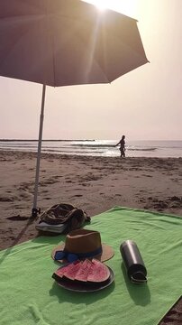 Vertical video of a beach towel spread on golden sand with freshly sliced watermelon on a plate, a sun hat with sunglasses, and a reusable water bottle. In the distance, a woman enjoys the sea